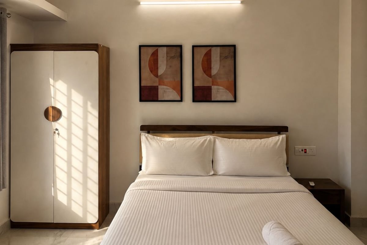 A hotel room with a bed, a ceiling fan, two wall-mounted artworks, a bedside table, and a door with a wooden frame.