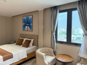 Une chambre d’hôtel avec un lit, une chaise, une petite table, un tableau et une fenêtre avec des rideaux.