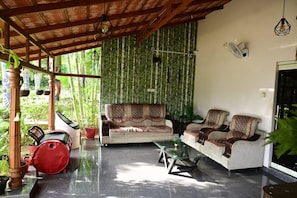 Un espacio de descanso al aire libre con techo, que incluye un sofá, sillones, una mesa de centro de vidrio y plantas en macetas.