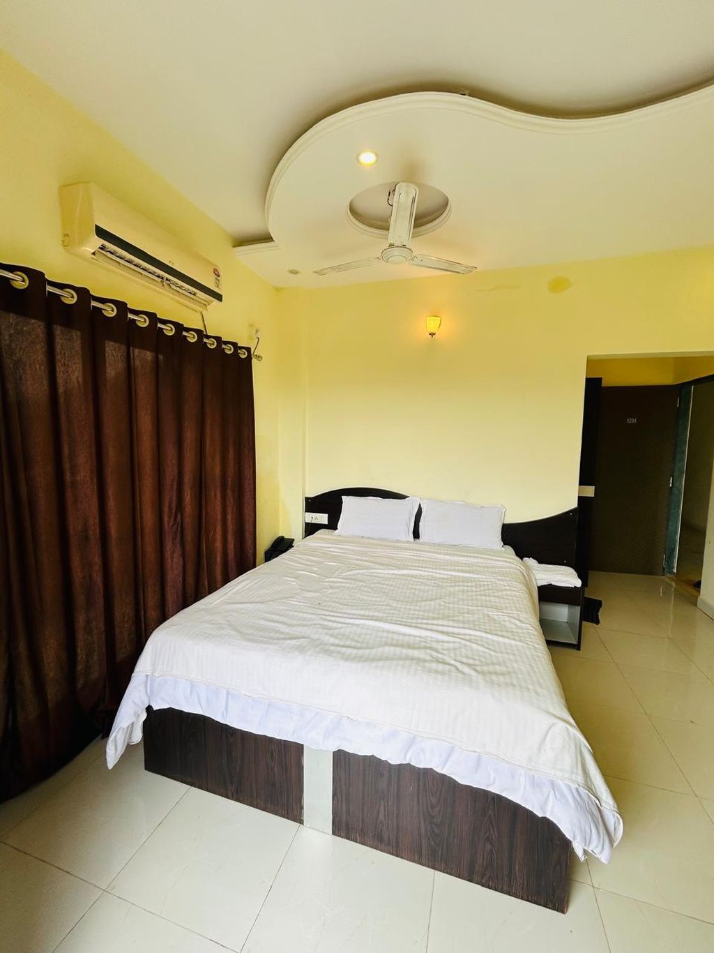 Habitación de hotel con una cama, ventilador de techo, equipo de aire acondicionado, mesita de noche y una puerta.