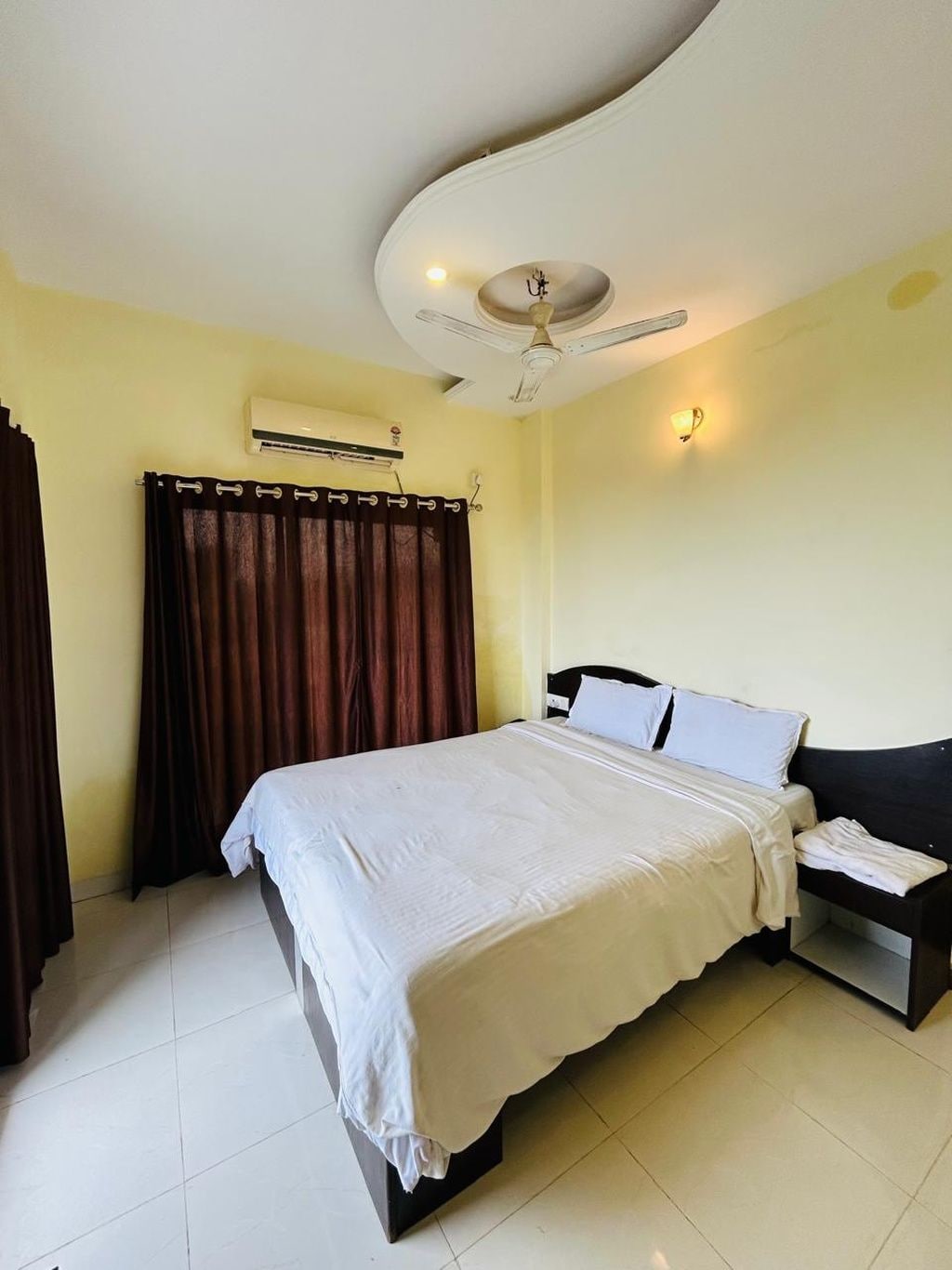 Habitación de hotel con cama, mesita de noche, ventilador de techo, aire acondicionado y cortina.