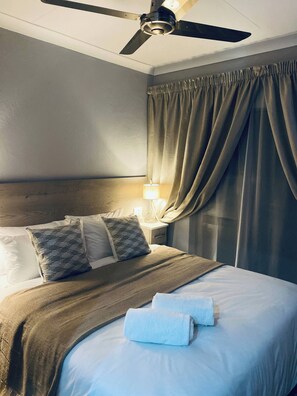 A hotel room with a bed, a ceiling fan, a nightstand with a lamp, and a window with curtains. - 23 on Jupiter Polokwane Unit (Polokwane)