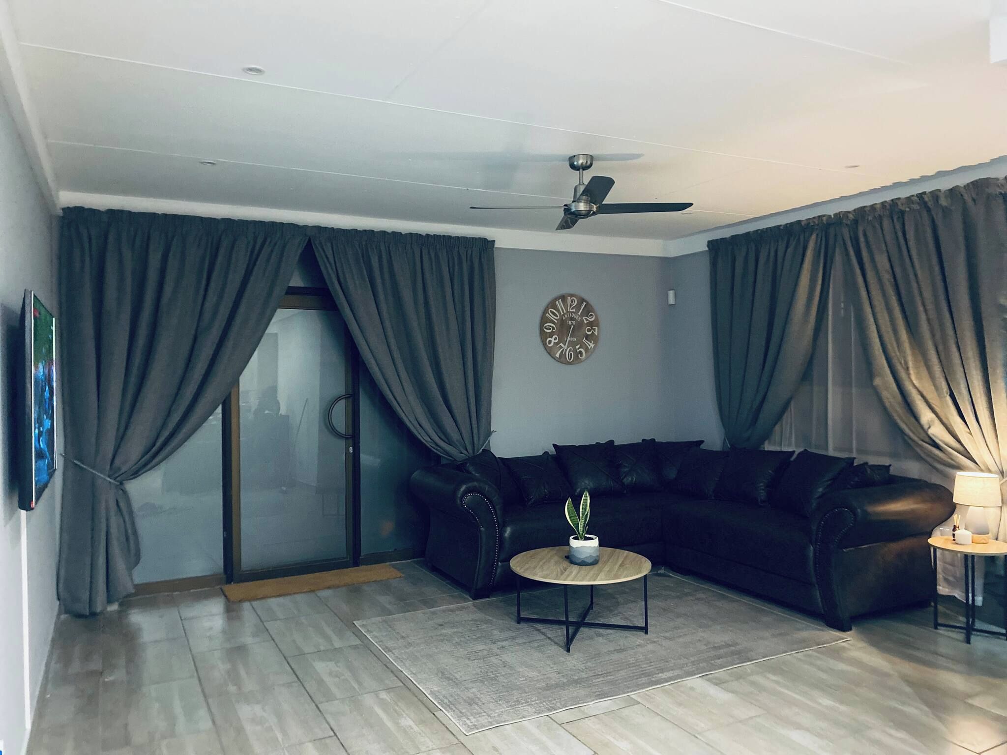 A living room with a black leather sofa, a round coffee table, a potted plant, a wall clock, and a ceiling fan.