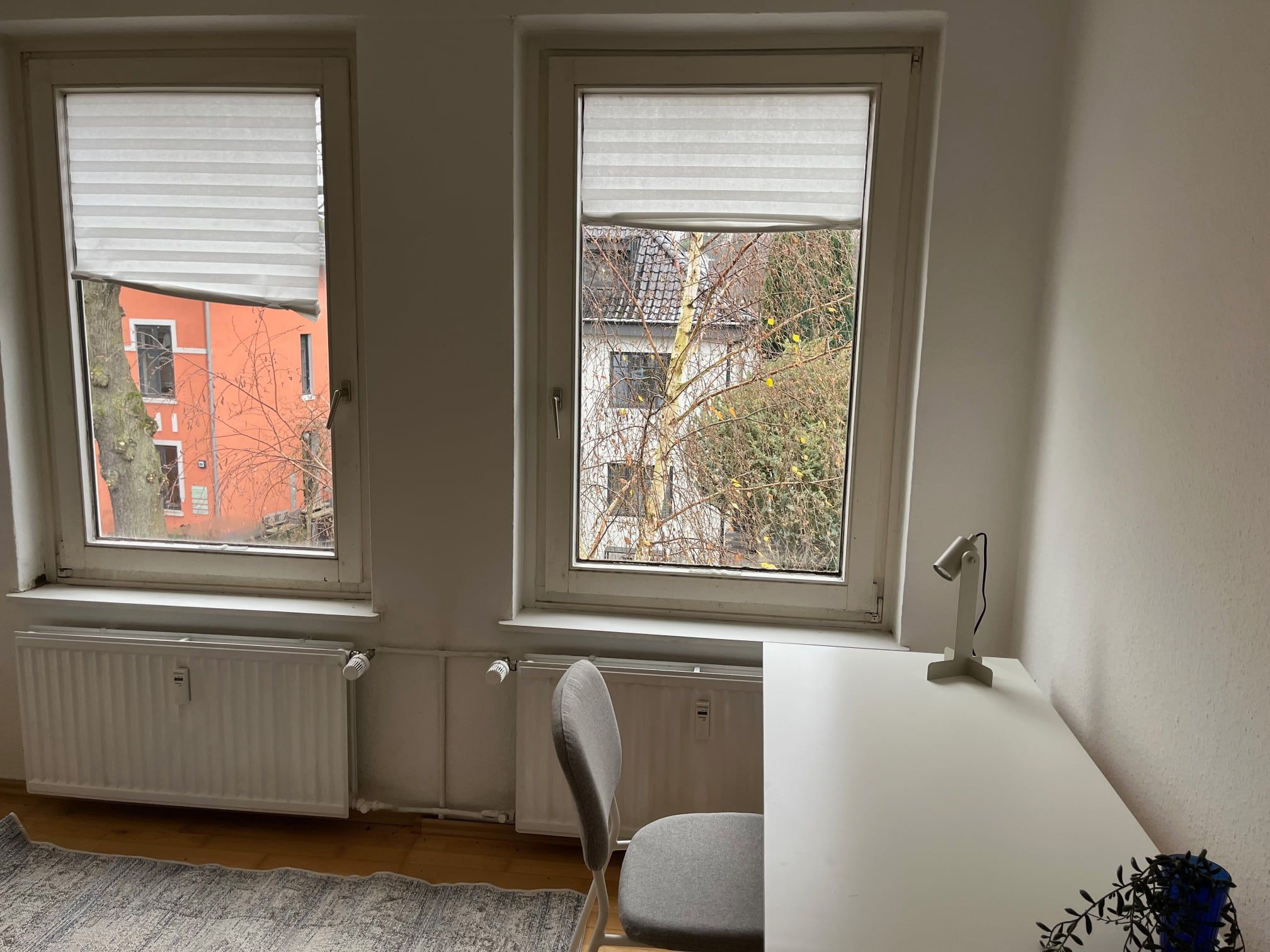 Ein Zimmer mit zwei Fenstern, einem Heizkörper, einem Stuhl und einem Schreibtisch.