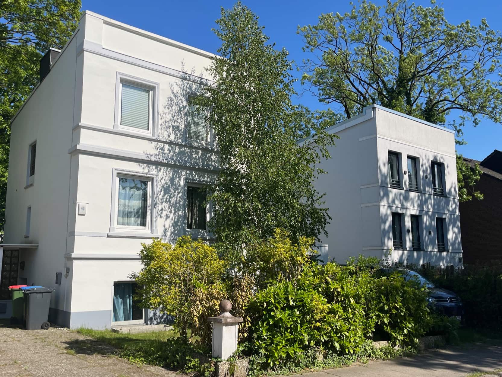 Ein zweistöckiges Gebäude mit flacher Fassade, zahlreichen Fenstern und einem kleinen Garten mit Baum und Sträuchern.