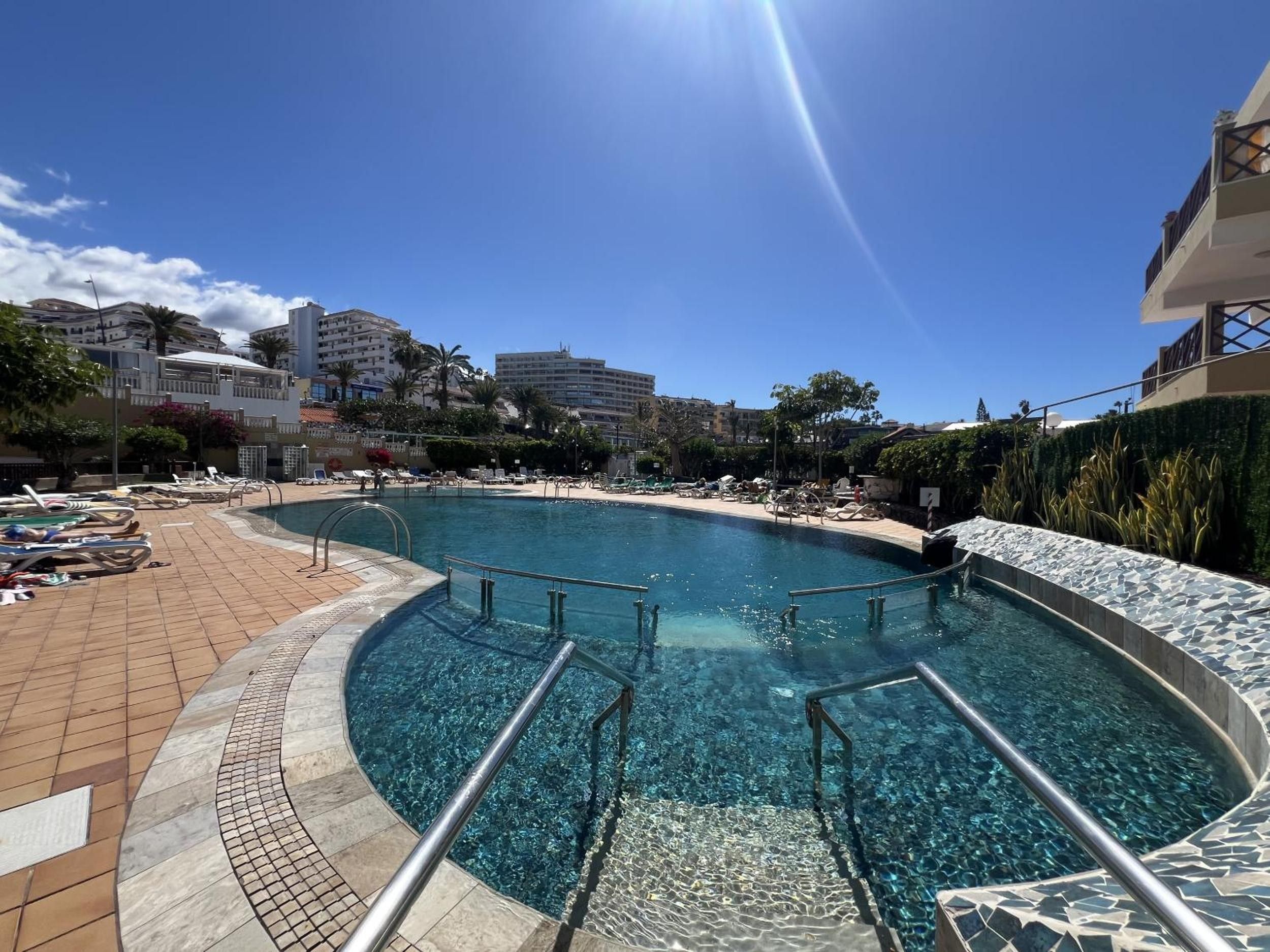 A clear blue swimming pool with a tiled deck, surrounded by lounge chairs and a sunshade.