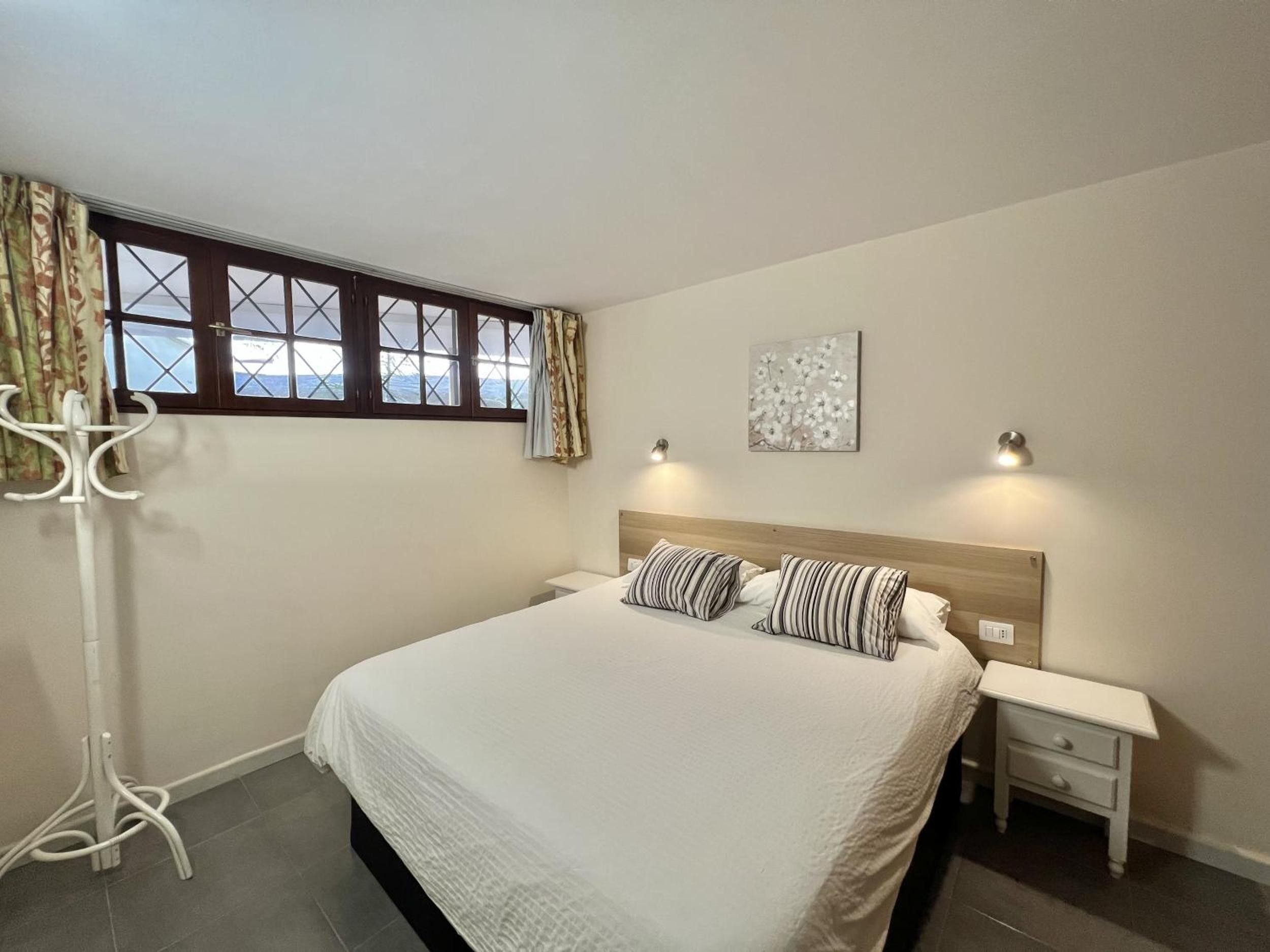 A bedroom with a bed, two bedside tables, a nightstand, a window with curtains, and a wall-mounted light fixture.