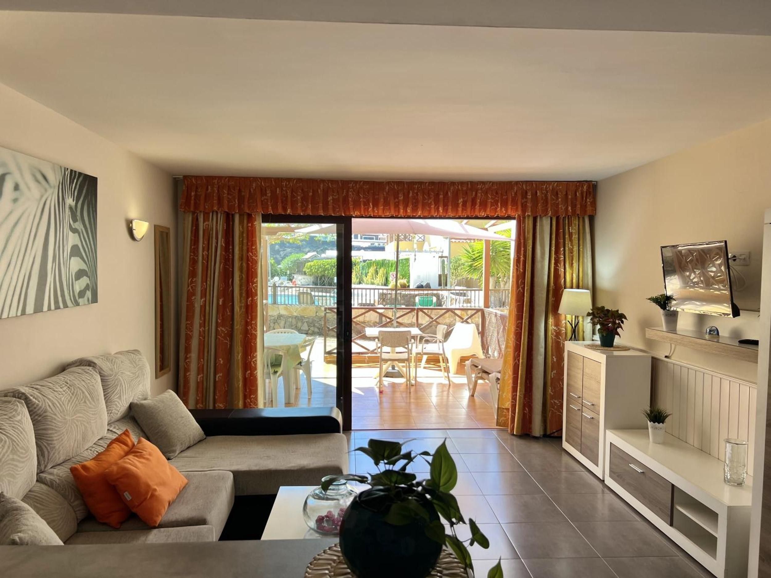 A living room with a sofa, coffee table, and TV stand. There is a sliding glass door leading to an outdoor patio with chairs and a pool.