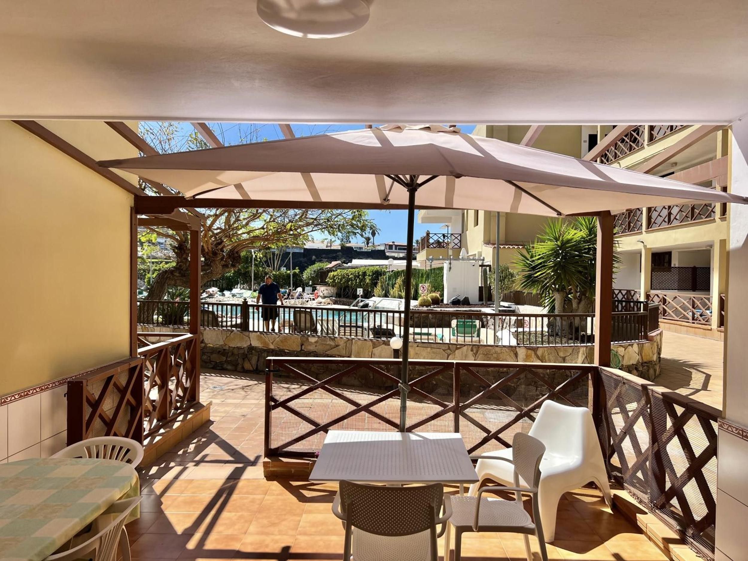 An outdoor patio with a table and chairs, a covered area, and a view of a pool and buildings in the background.