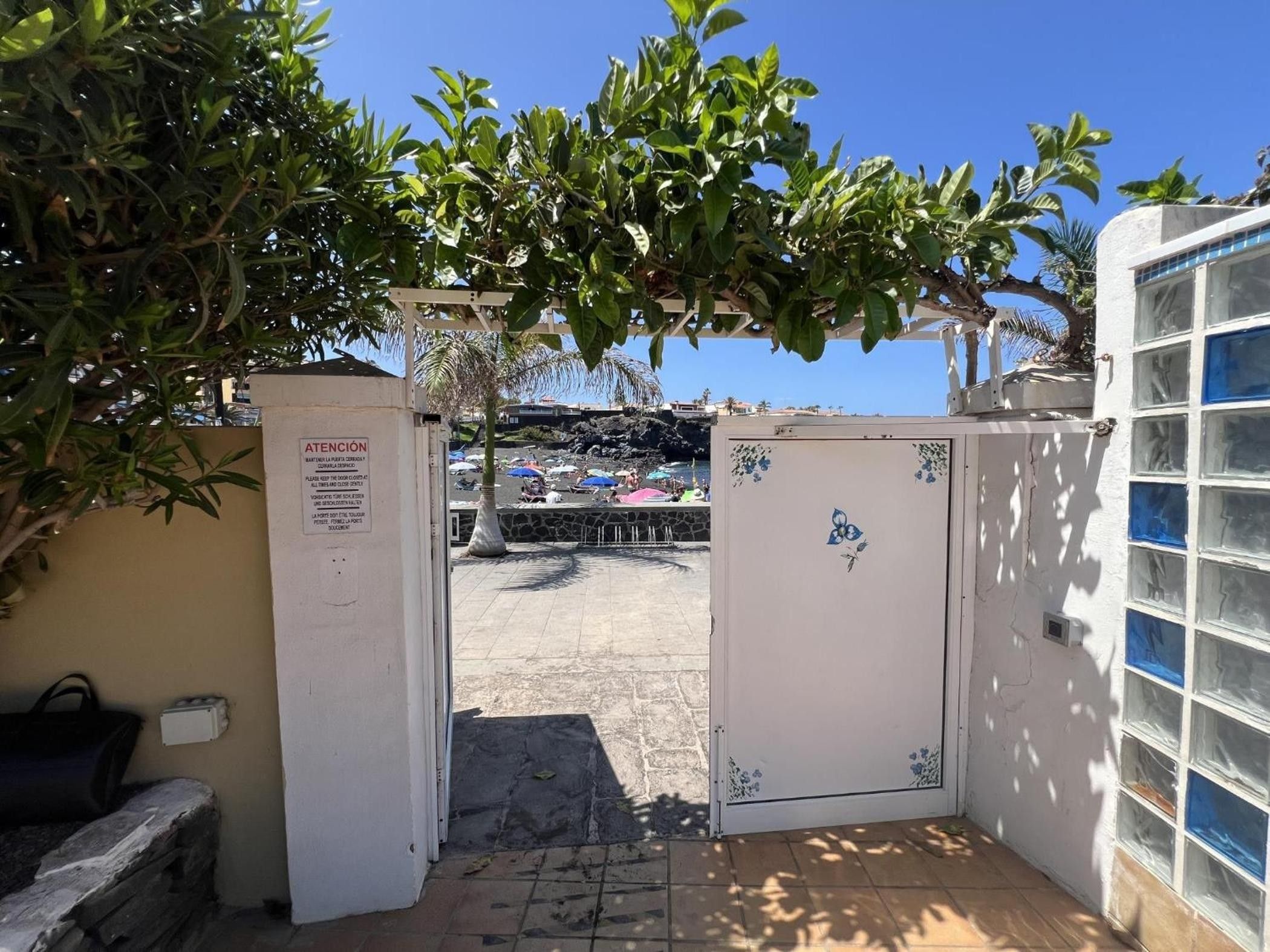 A gated entrance with a sign, a stone pathway, and a view of a beach with umbrellas.