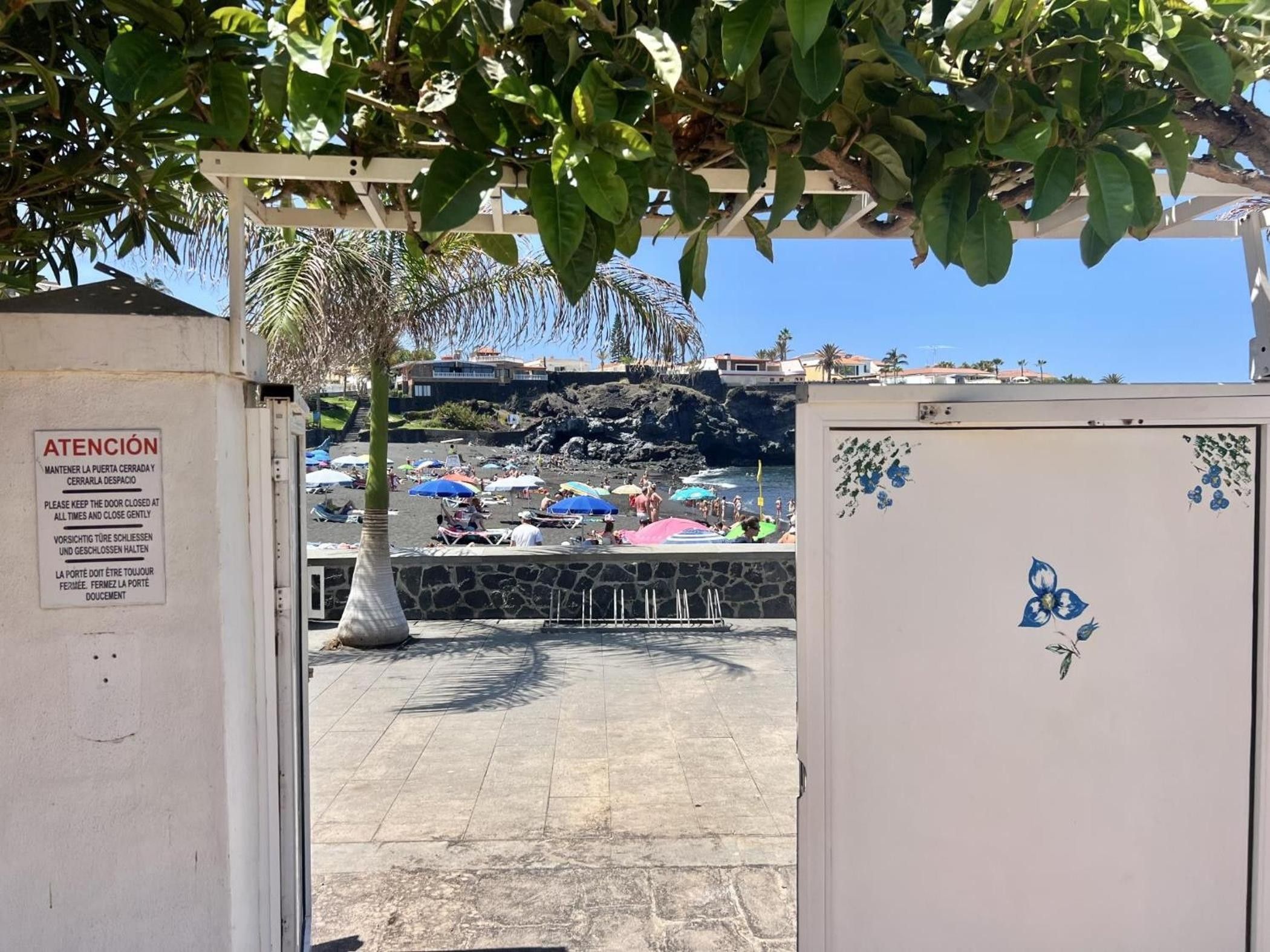 A beachside entrance with a sign instructing to keep the door closed and a view of the beach with umbrellas and people.