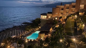 A coastal hotel with a swimming pool, beachfront umbrellas, and a view of the sea.