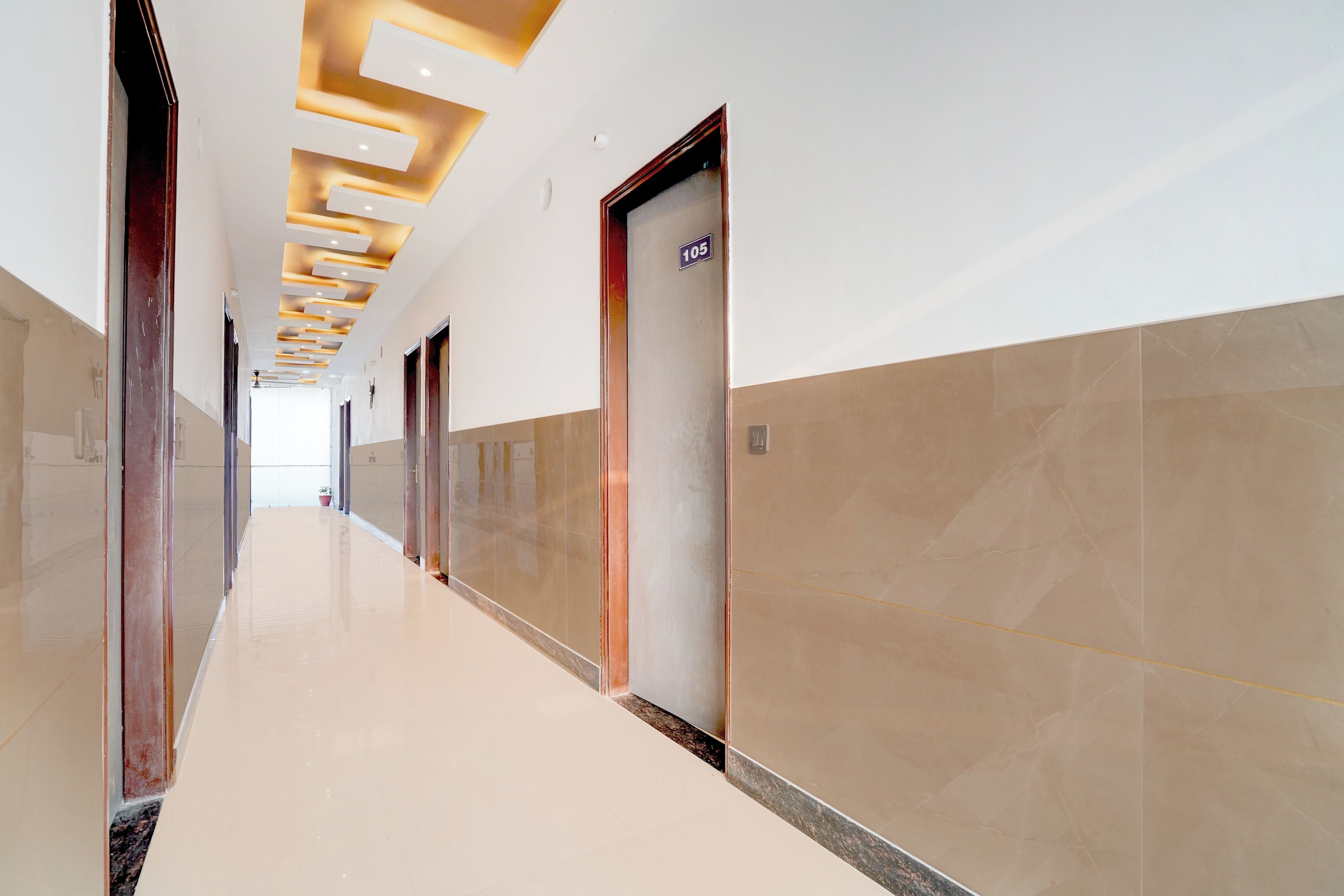 A long, narrow hotel corridor with a modern ceiling design, doors on both sides, and a tiled floor.