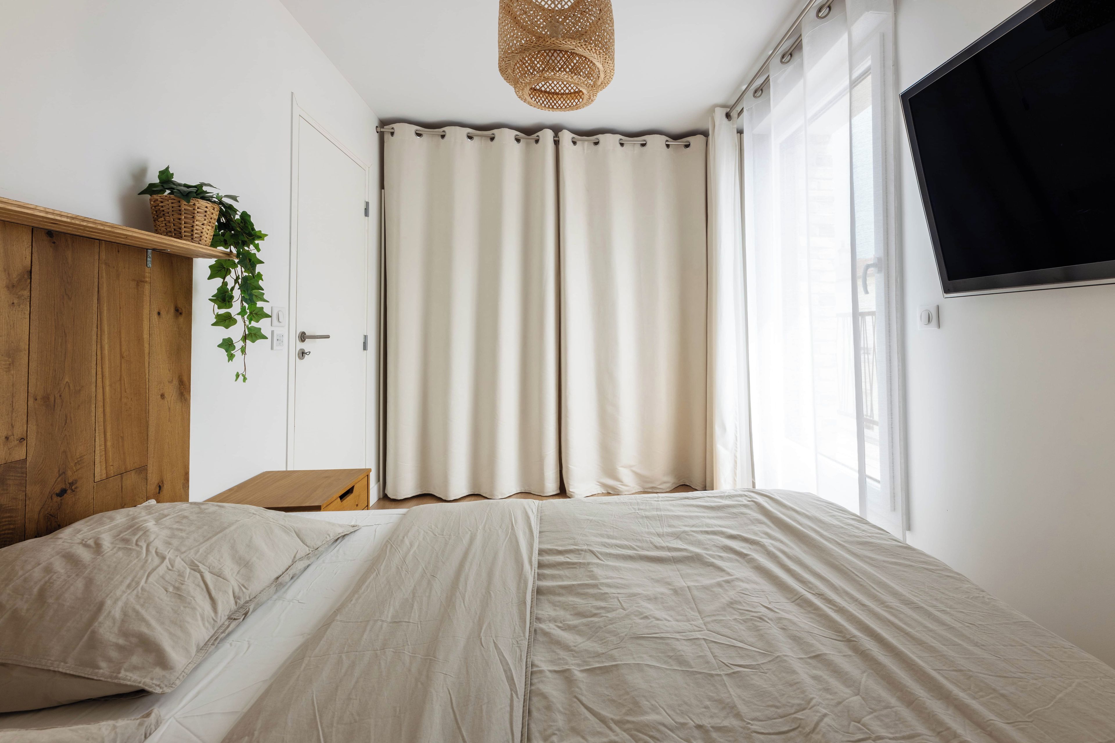 Un dormitorio con cama, cabecera de madera, televisor montado en la pared y ventana con cortinas.