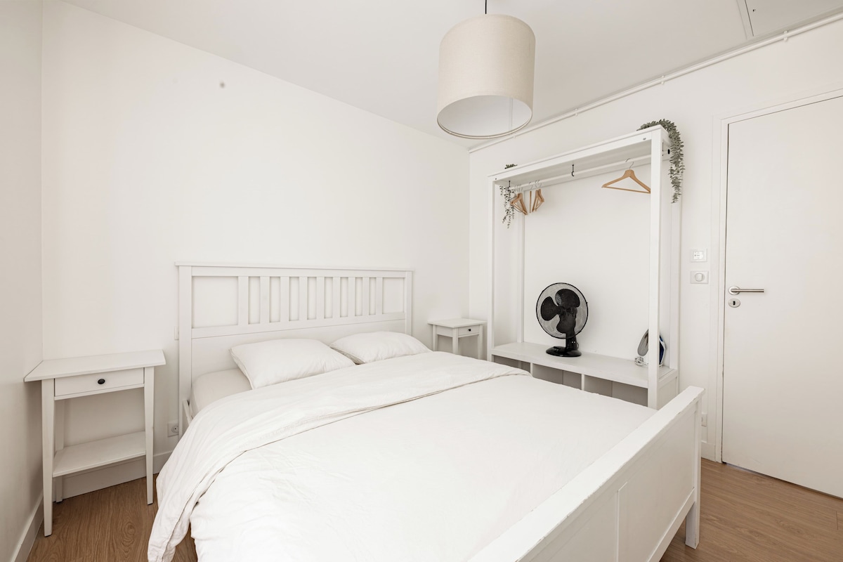 Un dormitorio blanco con una cama, mesitas de noche, un espejo, un ventilador y una puerta.