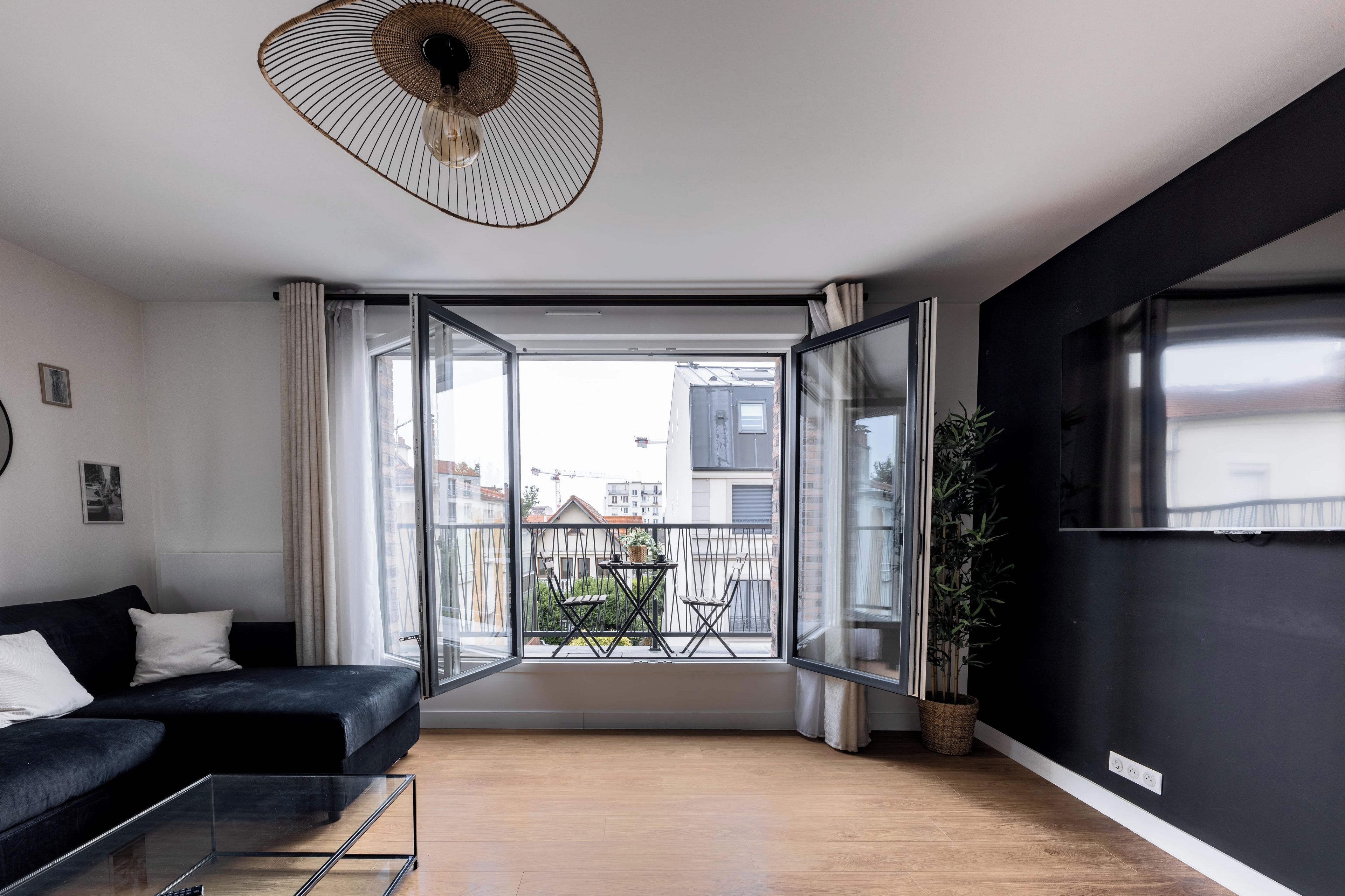 Een moderne woonkamer met een zwarte bank, een glazen salontafel en uitzicht op een balkon met een hekwerk en plantenpotten.