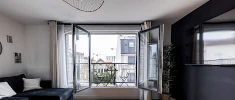 Un salon moderne avec un canapé noir, une table basse en verre et une vue sur un balcon doté d’une balustrade et de plantes en pot.