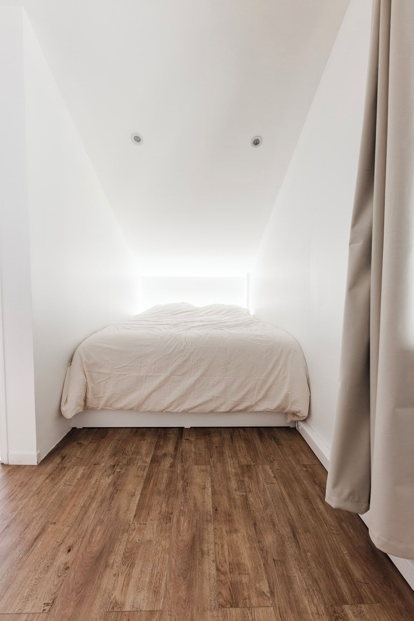Een slaapkamer met een schuine kap, een bed met een witte dekbedovertrek, houten vloeren en een raam met gordijnen.