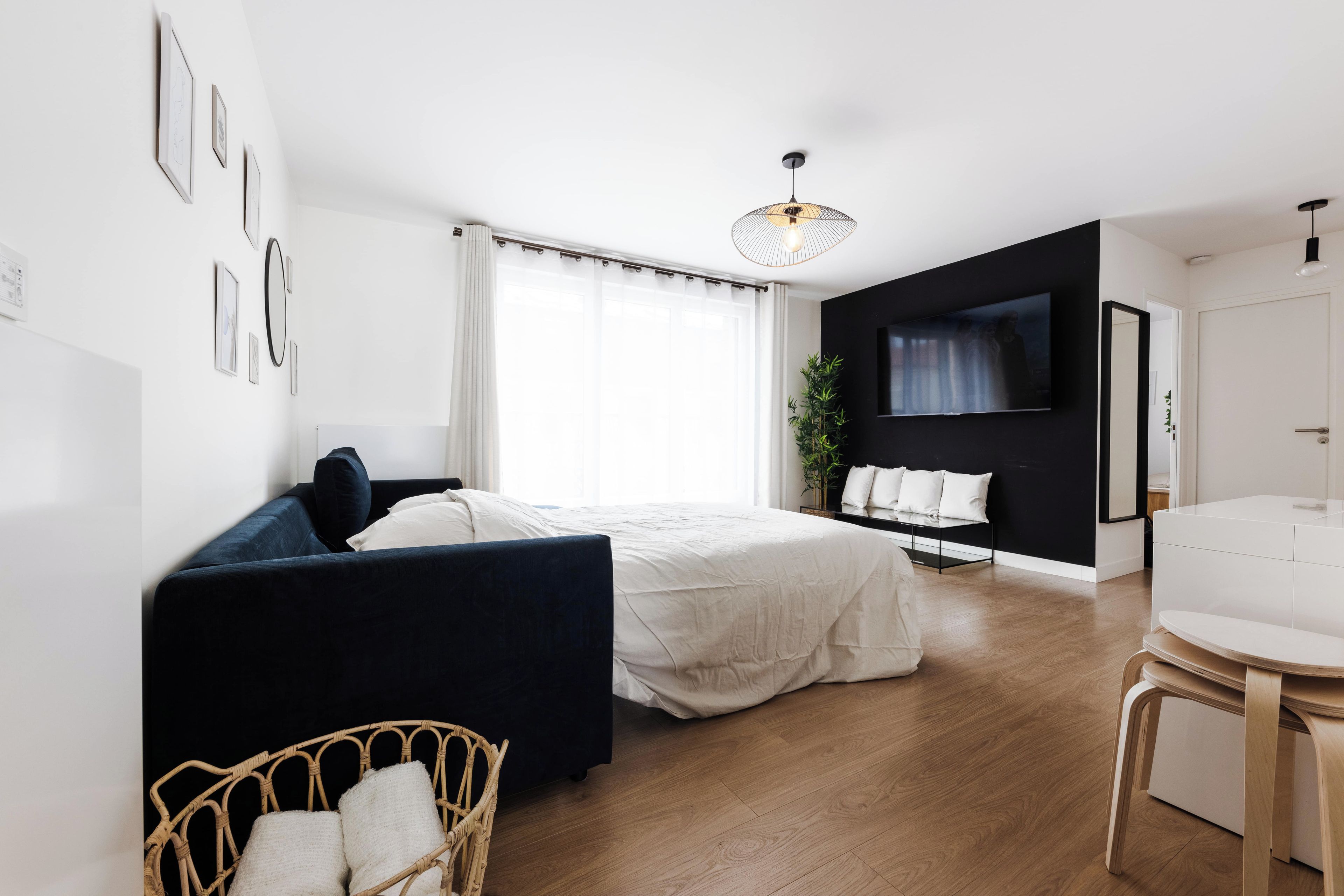 A modern bedroom with a bed, a dark sofa, a wicker chair, a bedside table, a TV, and a plant.