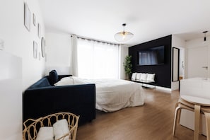 Une chambre à coucher moderne avec un lit, un canapé foncé, un fauteuil en osier, une table de chevet, un téléviseur et une plante.