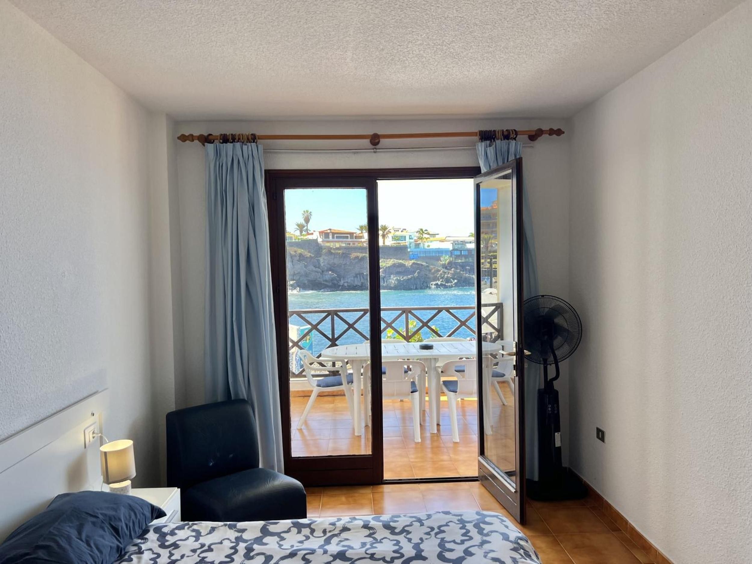 A bedroom with a bed, a chair, a table, and a view of the sea through a sliding glass door.
