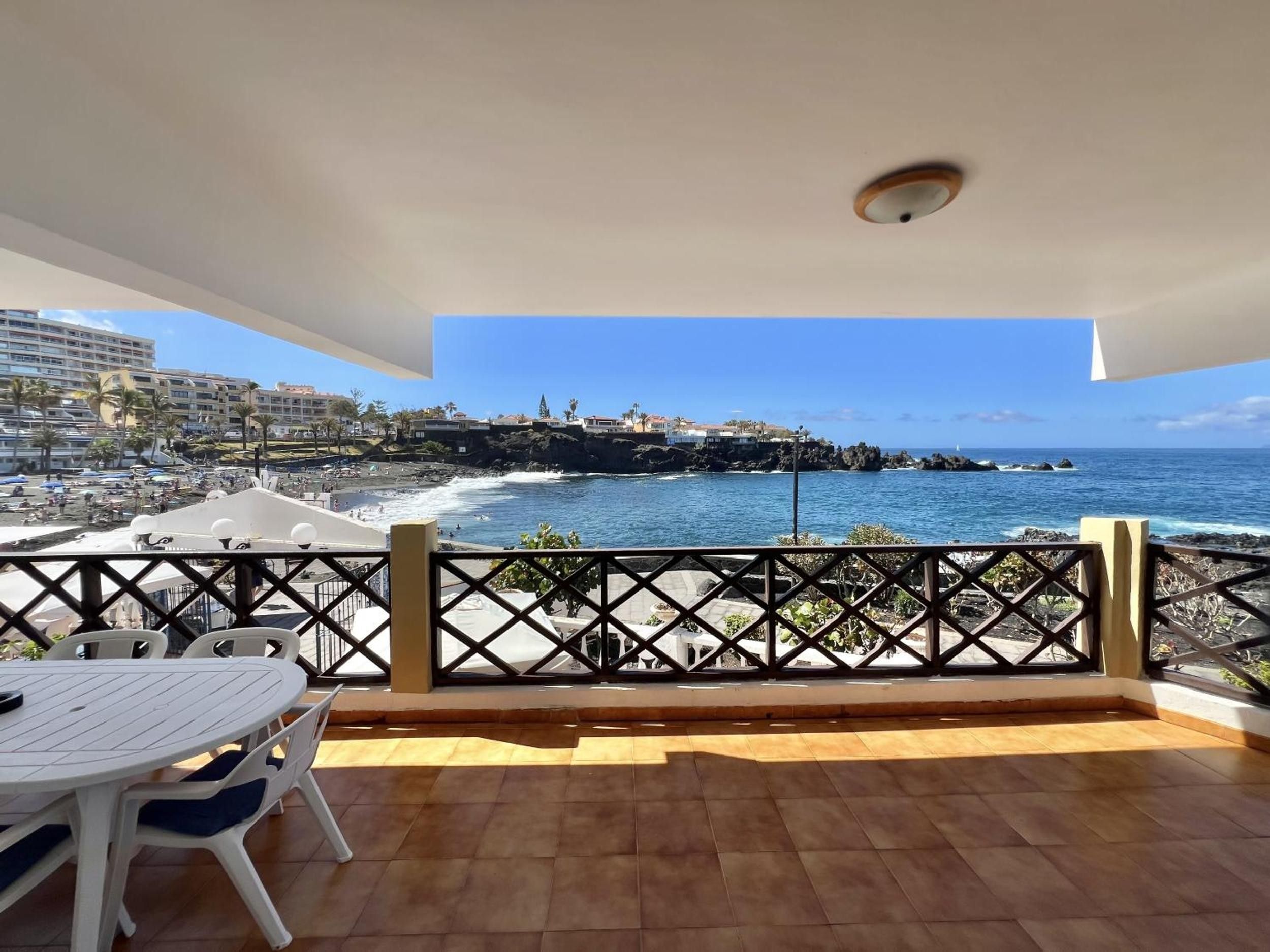 A balcony with a view of the ocean and a beach, a table with chairs, and a railing.