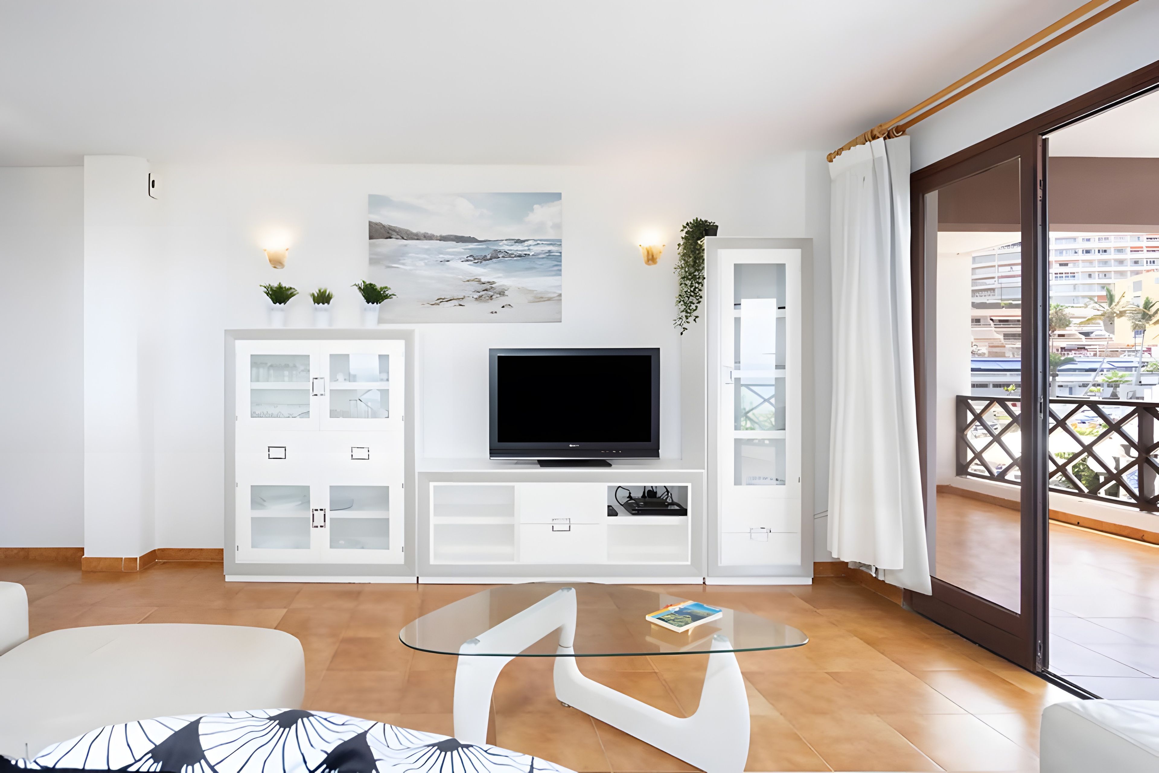 A modern living room with a flat-screen TV, a glass coffee table, and a view of the city through large windows.