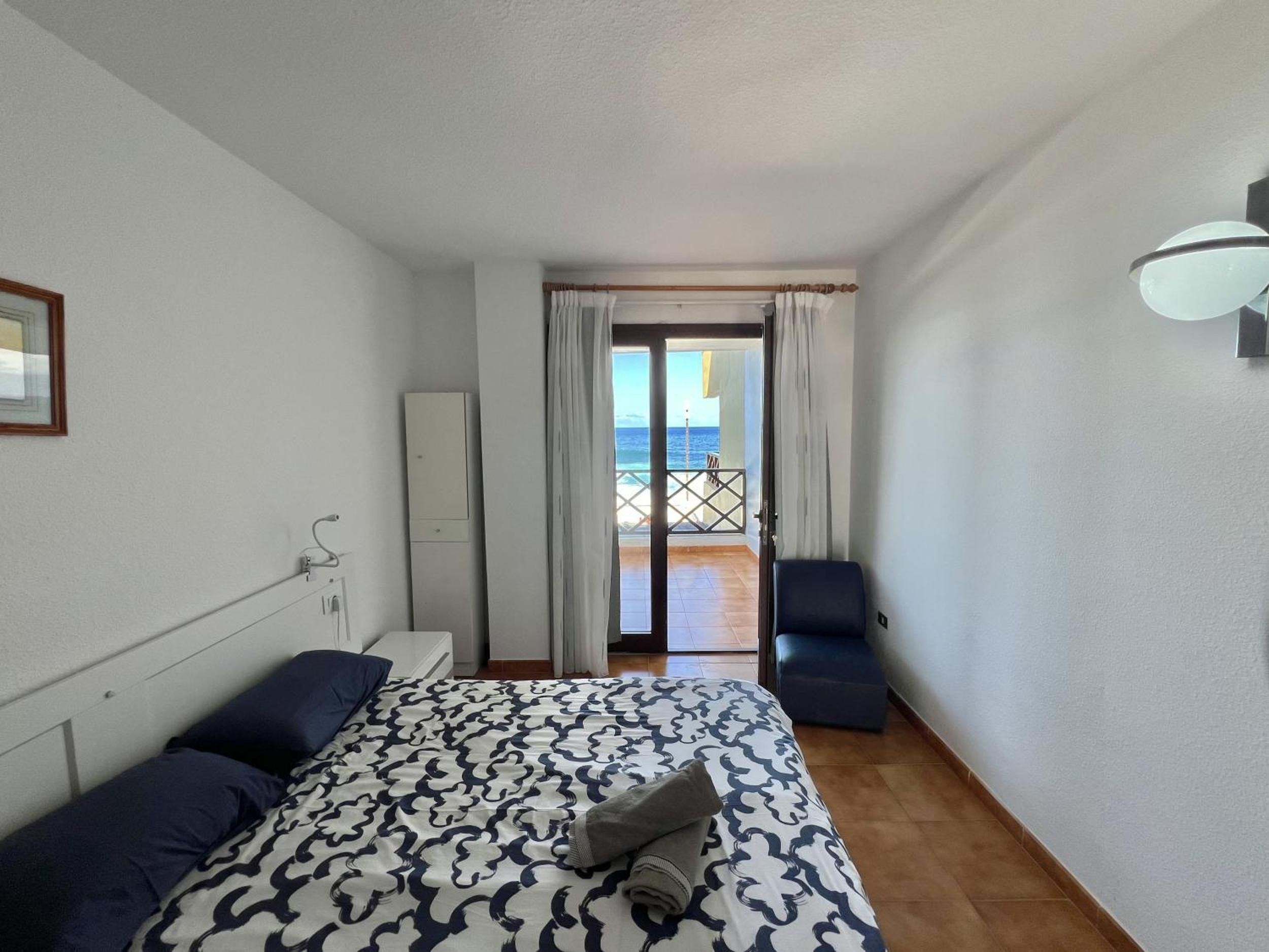 A hotel room with a bed, a chair, a wardrobe, and a view of the sea through an open door.