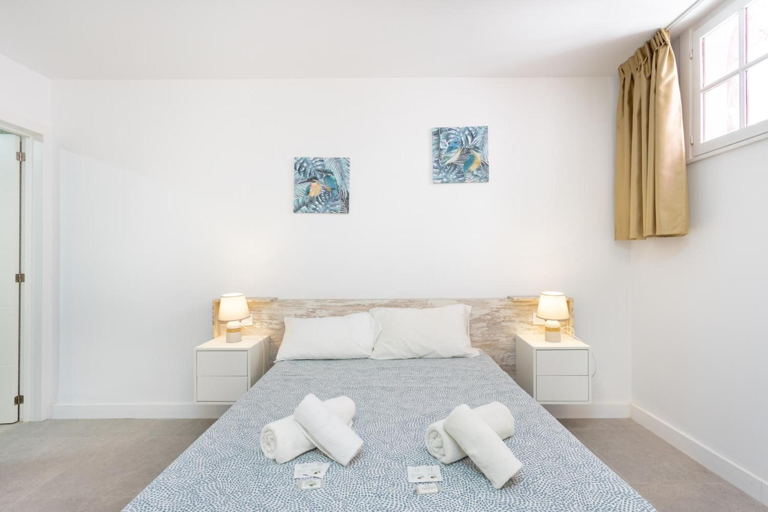 Un dormitorio con una cama, dos mesitas de noche con lámparas, un cabecero, dos obras de arte en la pared, una ventana con cortinas y toallas en el suelo.