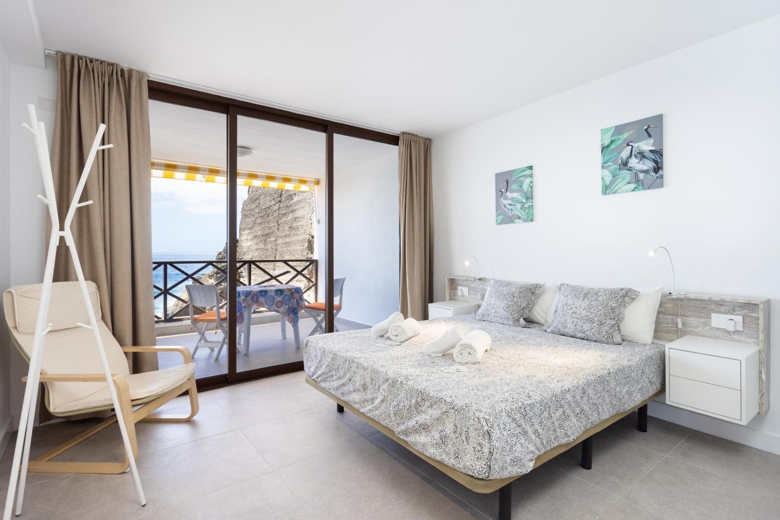Un dormitorio con cama, una silla, una mesita de noche y vista al mar a través de una puerta corrediza.