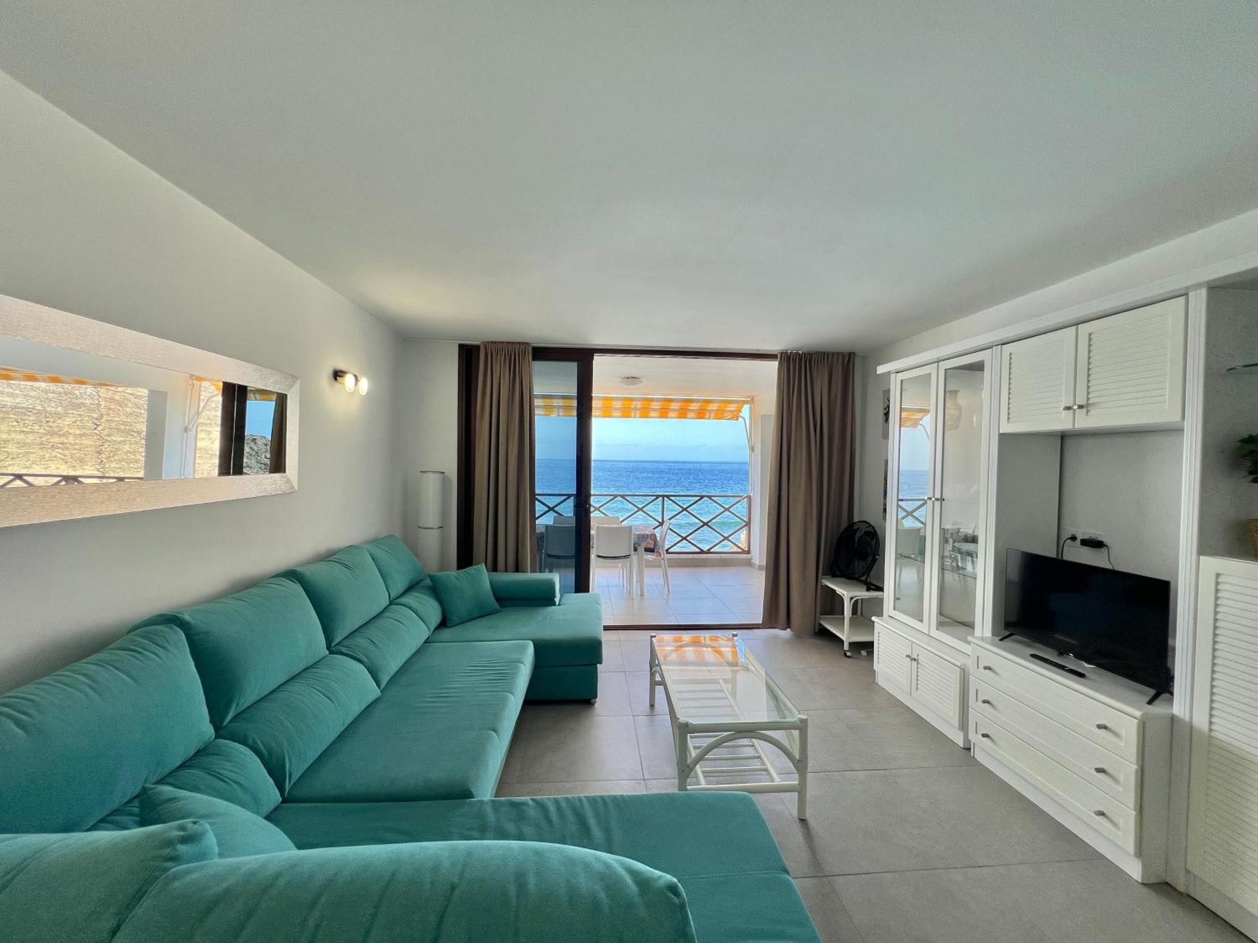 A modern living room with a sofa, a TV, and a balcony with a view of the sea.