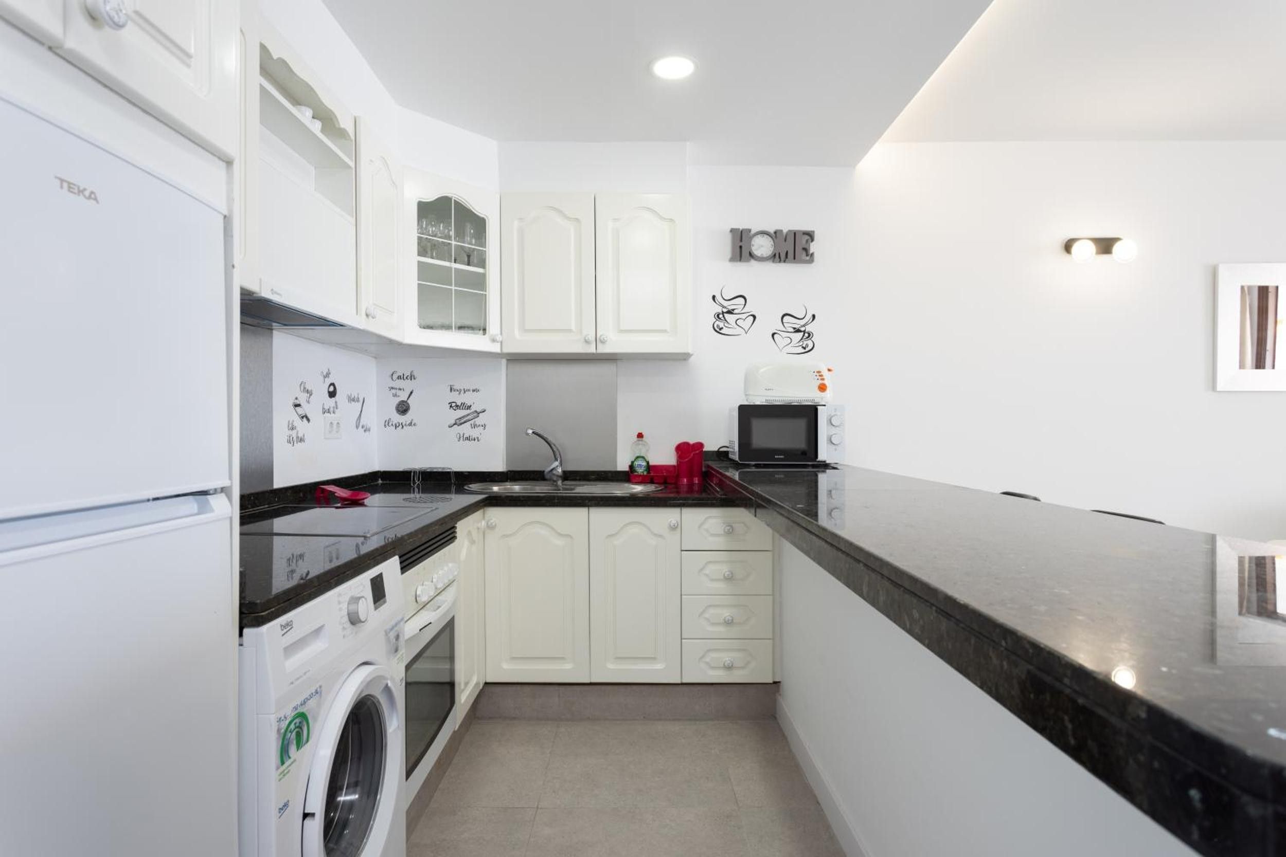 A compact kitchen with white cabinets, a black countertop, a microwave, a washing machine, and a sink.