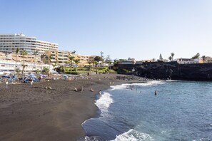 A beach with a hotel in the background, a rocky cliff, and palm trees.