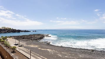 A coastal road with rocky terrain leading to the sea, a clear sky, and distant buildings.