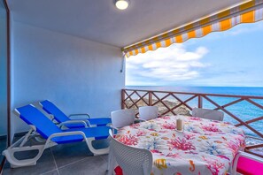 Un balcón con dos sillones azules, una mesa con mantel floral y vista al mar.