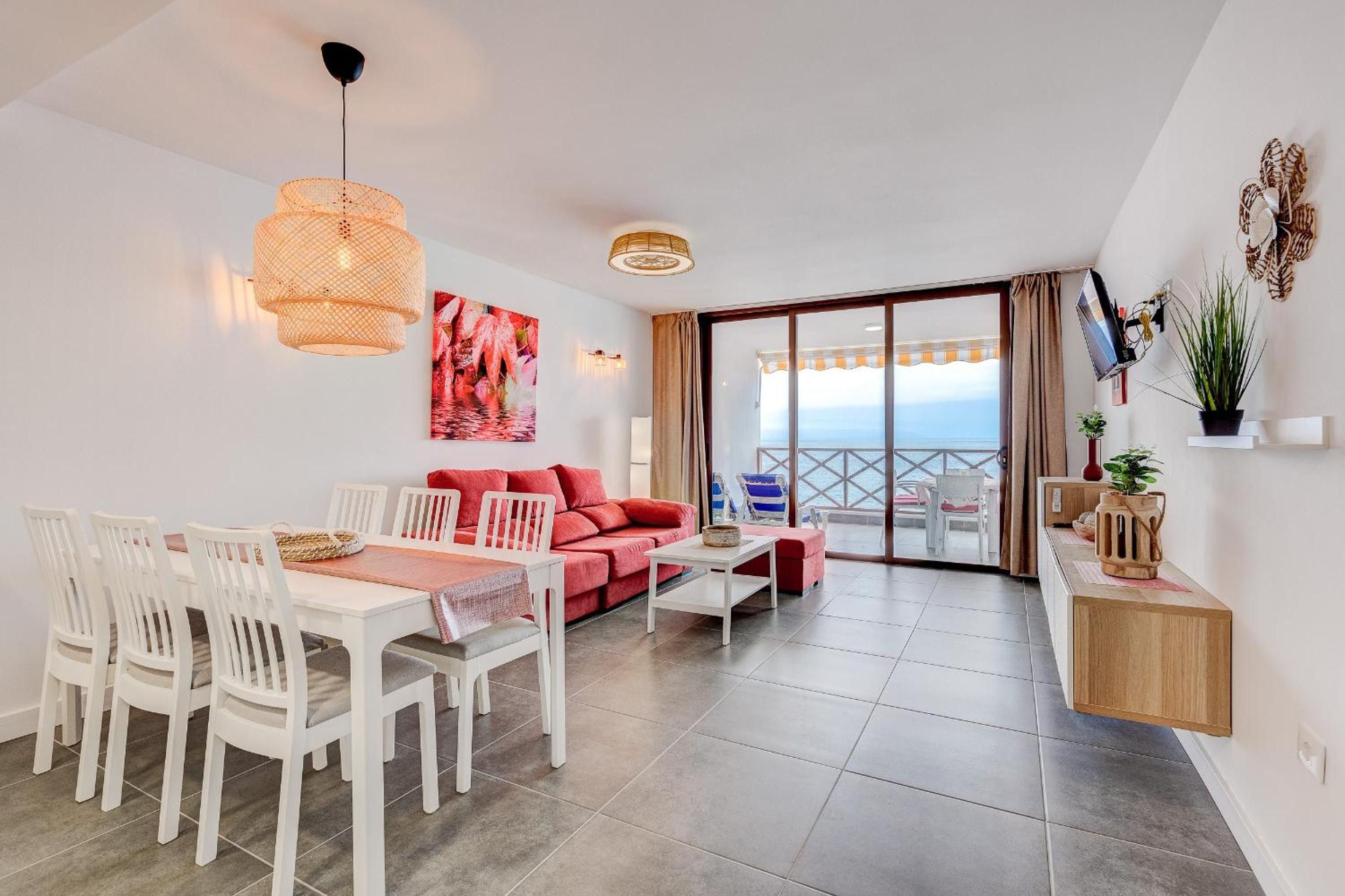 A modern living room with a dining area, a red sofa, white chairs, and a view of the sea through large windows.