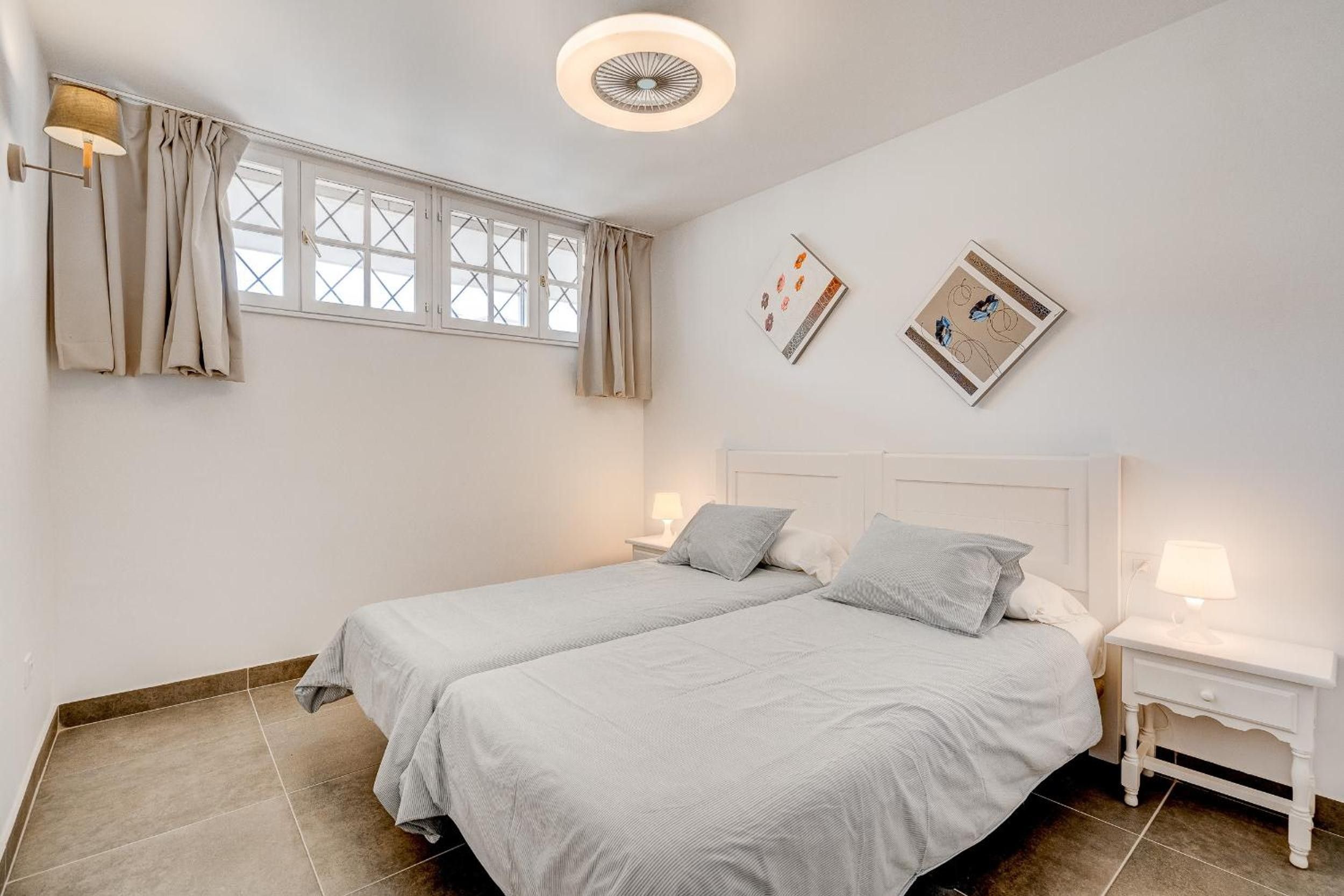 A bedroom with two beds, a window with curtains, a bedside table with a lamp, and a wall-mounted light fixture.