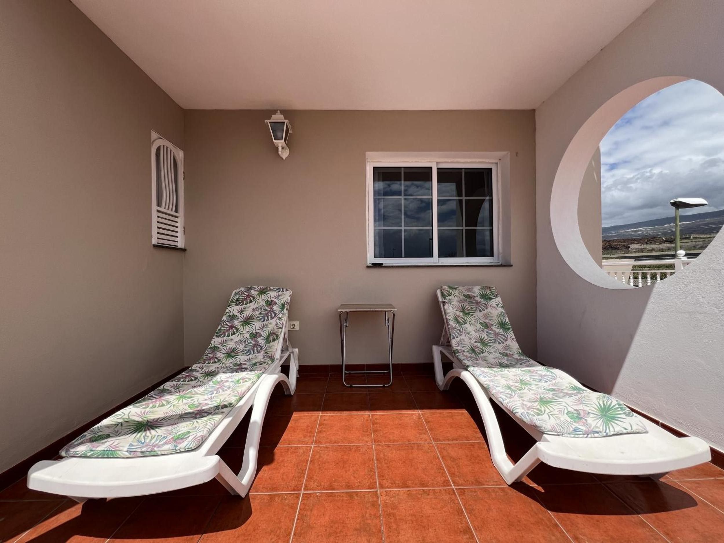 A balcony with two sun loungers, a small table, a window, and a wall-mounted light fixture.