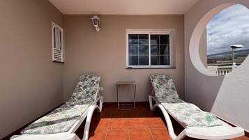 A balcony with two sun loungers, a small table, a window, and a wall-mounted light fixture.