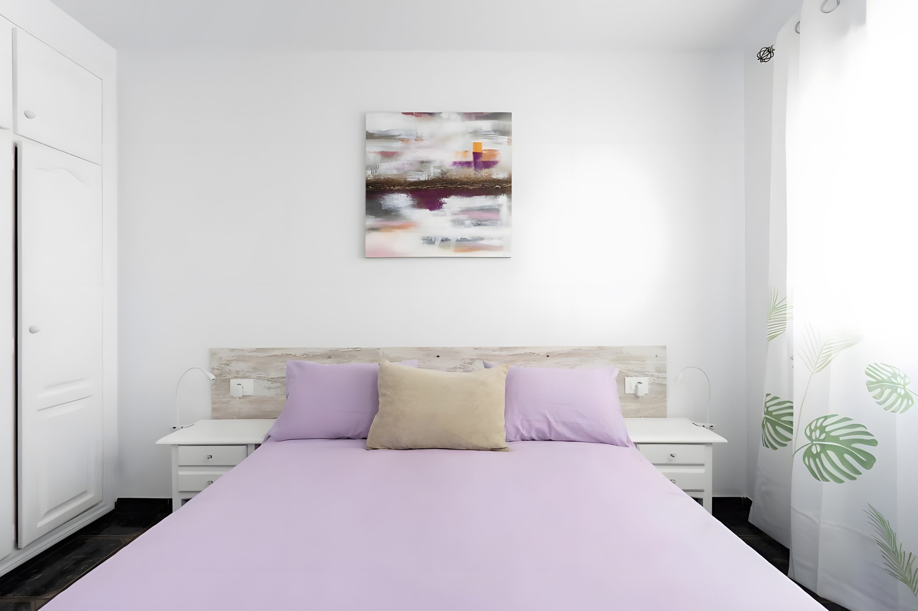 A bedroom with a bed, a nightstand, a wall-mounted artwork, and a plant.