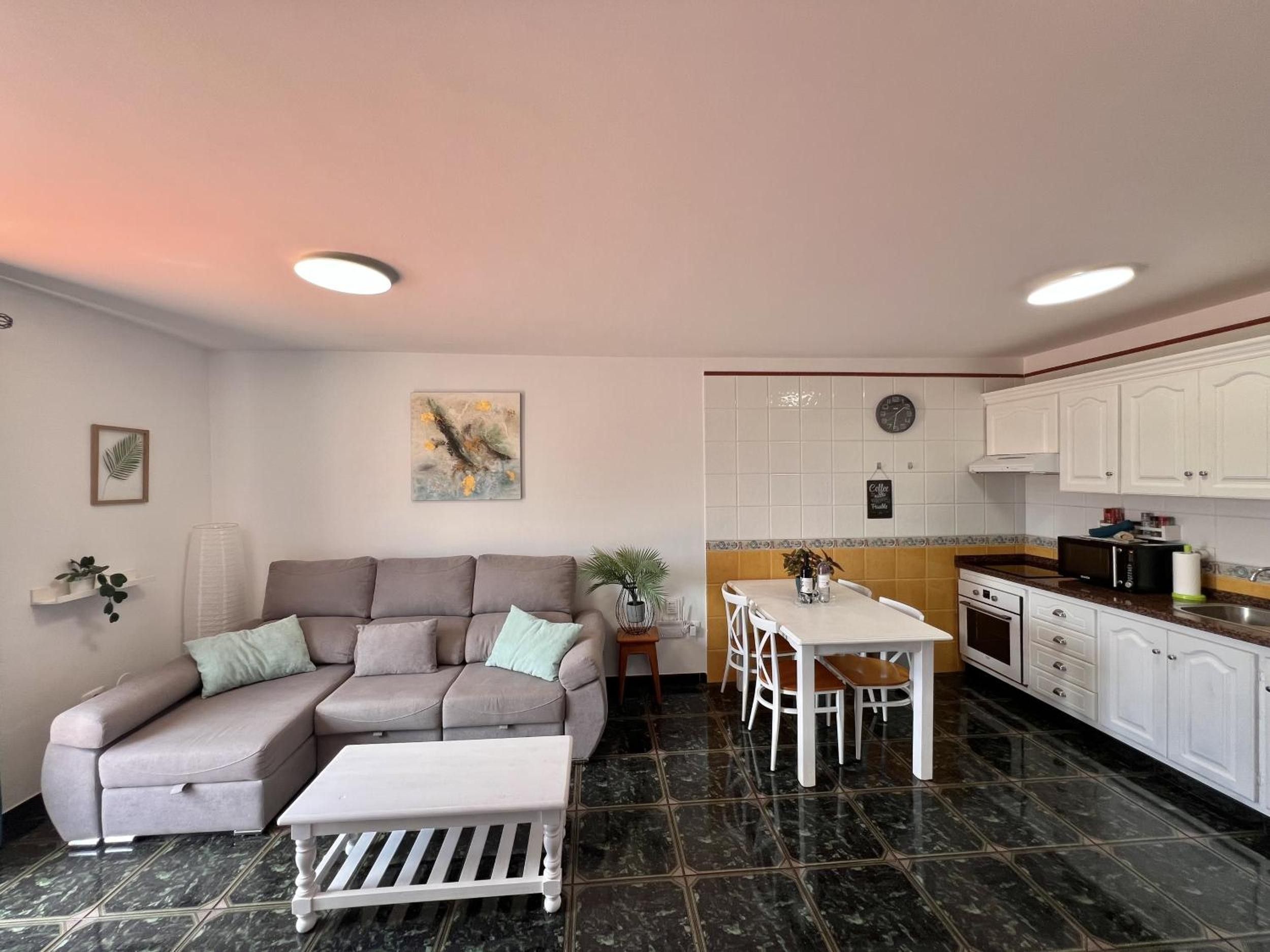 A living room with a sofa, a coffee table, and a dining area with a table and chairs. There is a kitchen with white cabinets and a microwave.