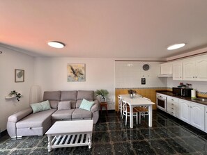 Un saló amb un sofà, una taula de centre i una zona de menjador amb taula i cadires. Hi ha una cuina amb armaris blancs i un microones.
