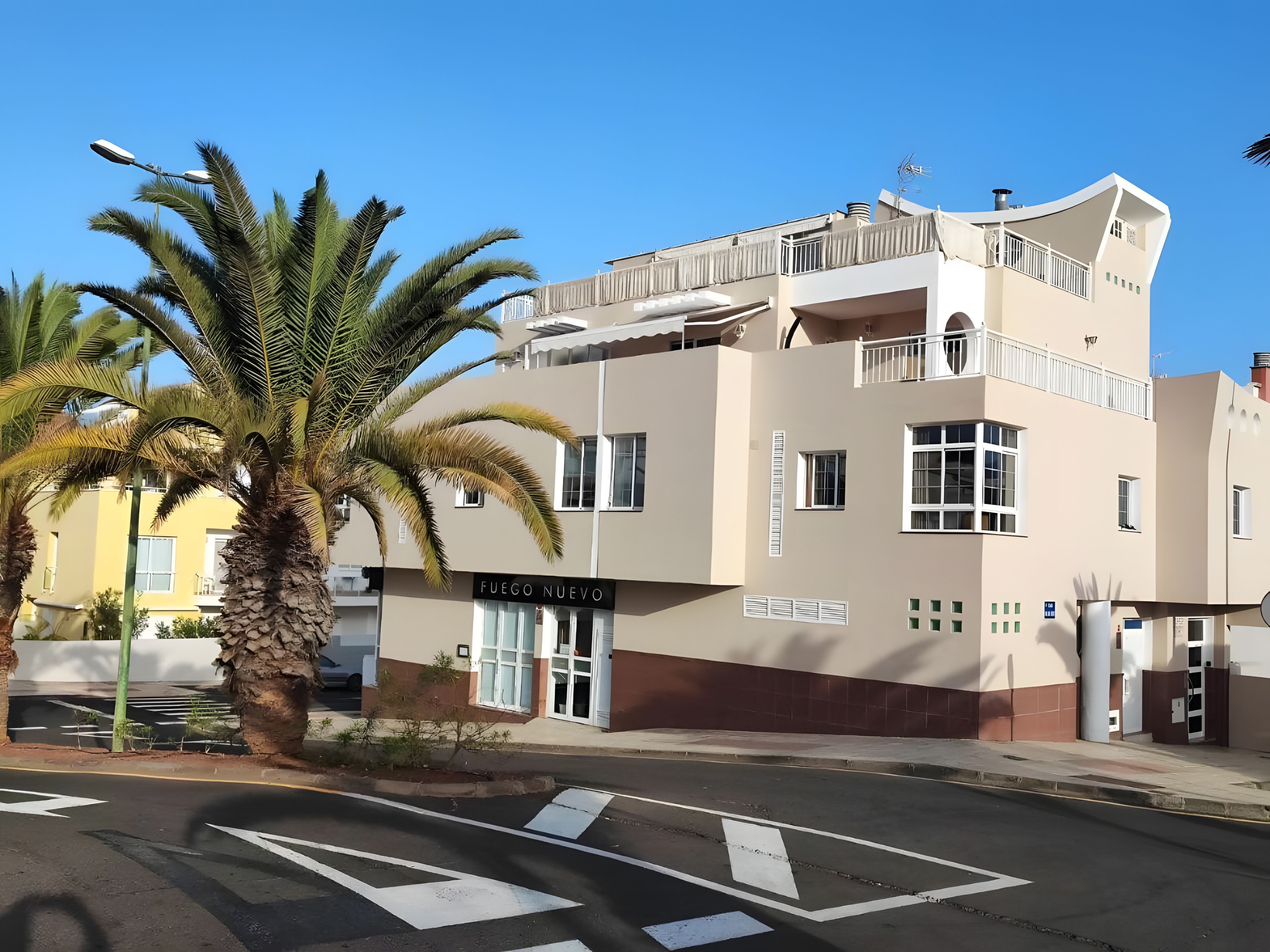 Un edificio de varios pisos con un cartel que dice 'FUEGO NUEVO' en la planta baja, un balcón en el nivel superior y una palmera en primer plano.