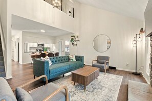 A spacious living area with a sofa, armchairs, a coffee table, and a rug. There is a kitchen in the background and a staircase leading to an upper level. - Cbus Hub · 2 Kings + Loft  2 twin beds · Sleeps 6 · Private Deck & Grill (Columbus)