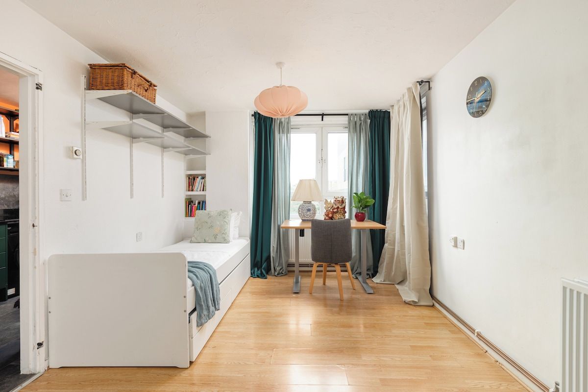 Une petite pièce comprenant un lit, un bureau et une chaise. On y trouve une fenêtre avec des rideaux, une horloge murale et une étagère avec un panier.