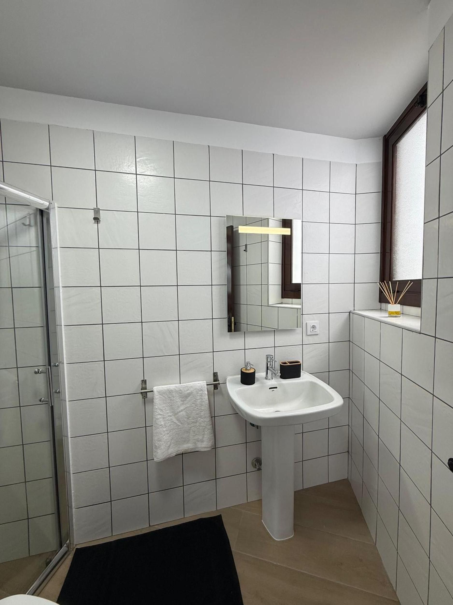 Ein Badezimmer mit weiß gefliesten Wänden, einem weißen Waschbecken, einem Spiegel und einer Duschkabine aus Glas.