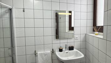 Ein Badezimmer mit weiß gefliesten Wänden, einem weißen Waschbecken, einem Spiegel und einer Duschkabine aus Glas.