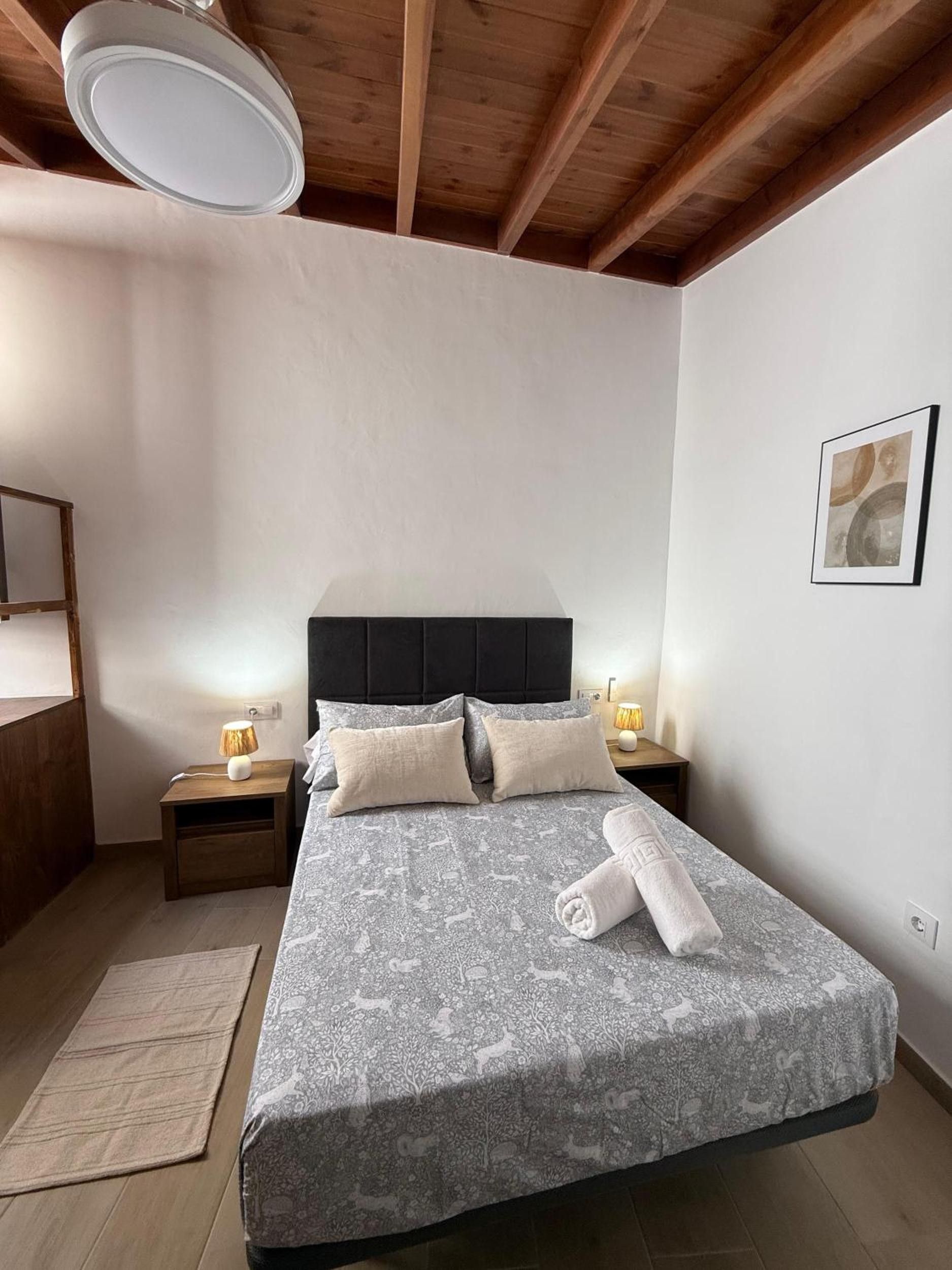 Un dormitorio con una cama, mesitas de noche, una lámpara, un cuadro enmarcado en la pared y techo de madera.
