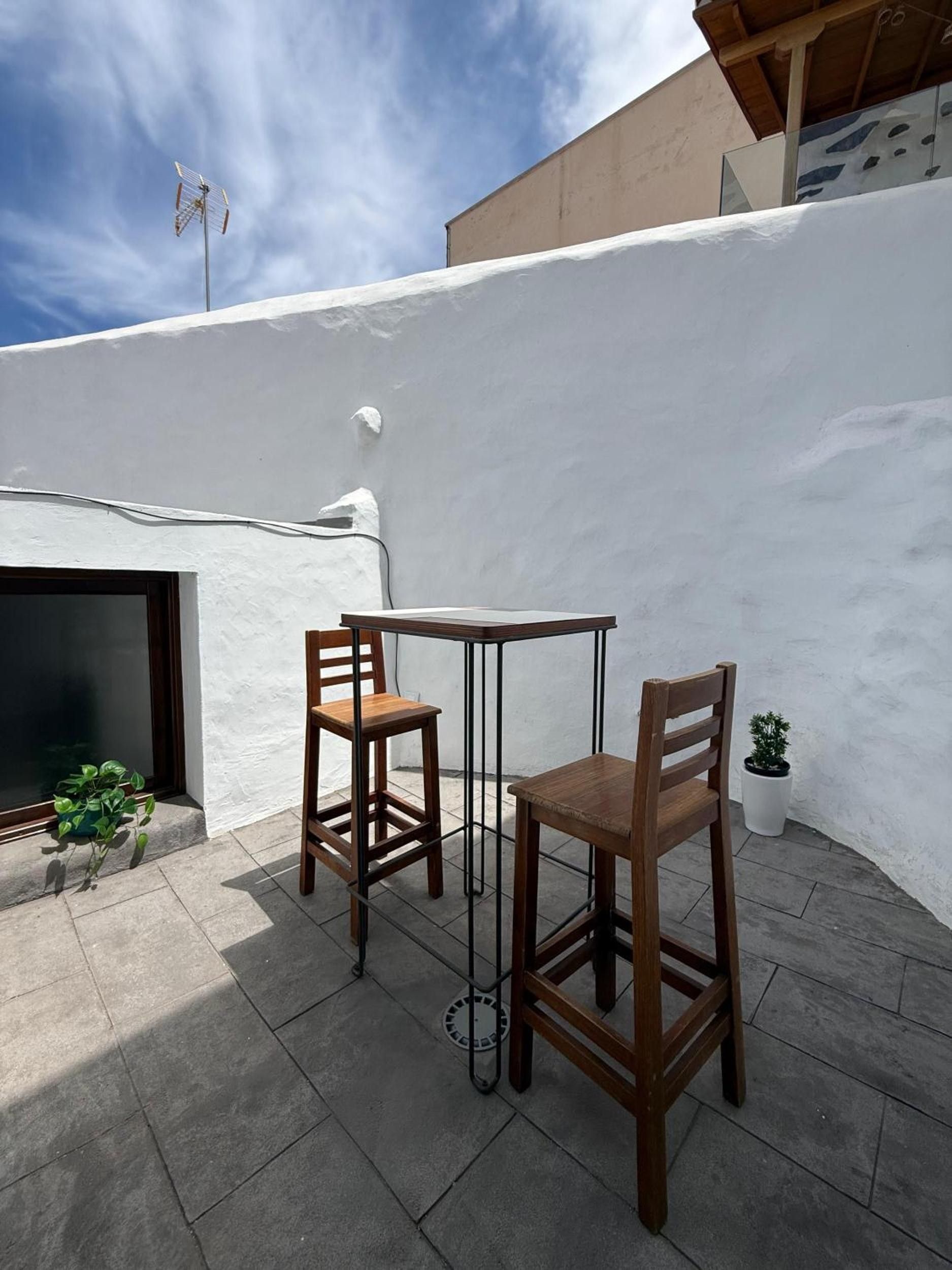 An outdoor patio with a table and two chairs, a potted plant, and a building with a white exterior.