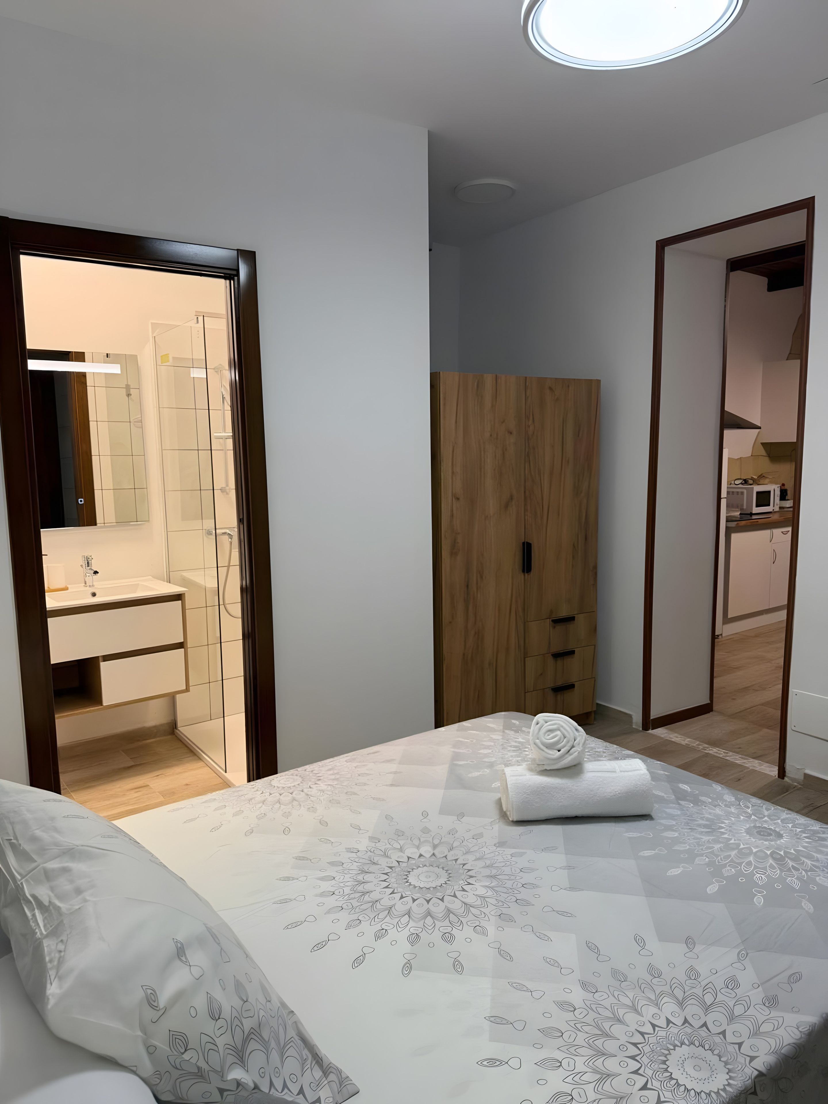 Een slaapkamer met een bed, een houten garderobe, een badkamer met een spiegel en wastafel, en een keuken die zichtbaar is door een open deur.